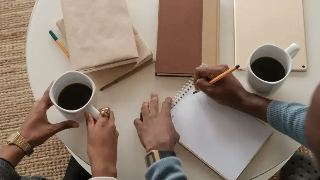Two people at a small round table with cups of coffee, one of them writing in a notebook.