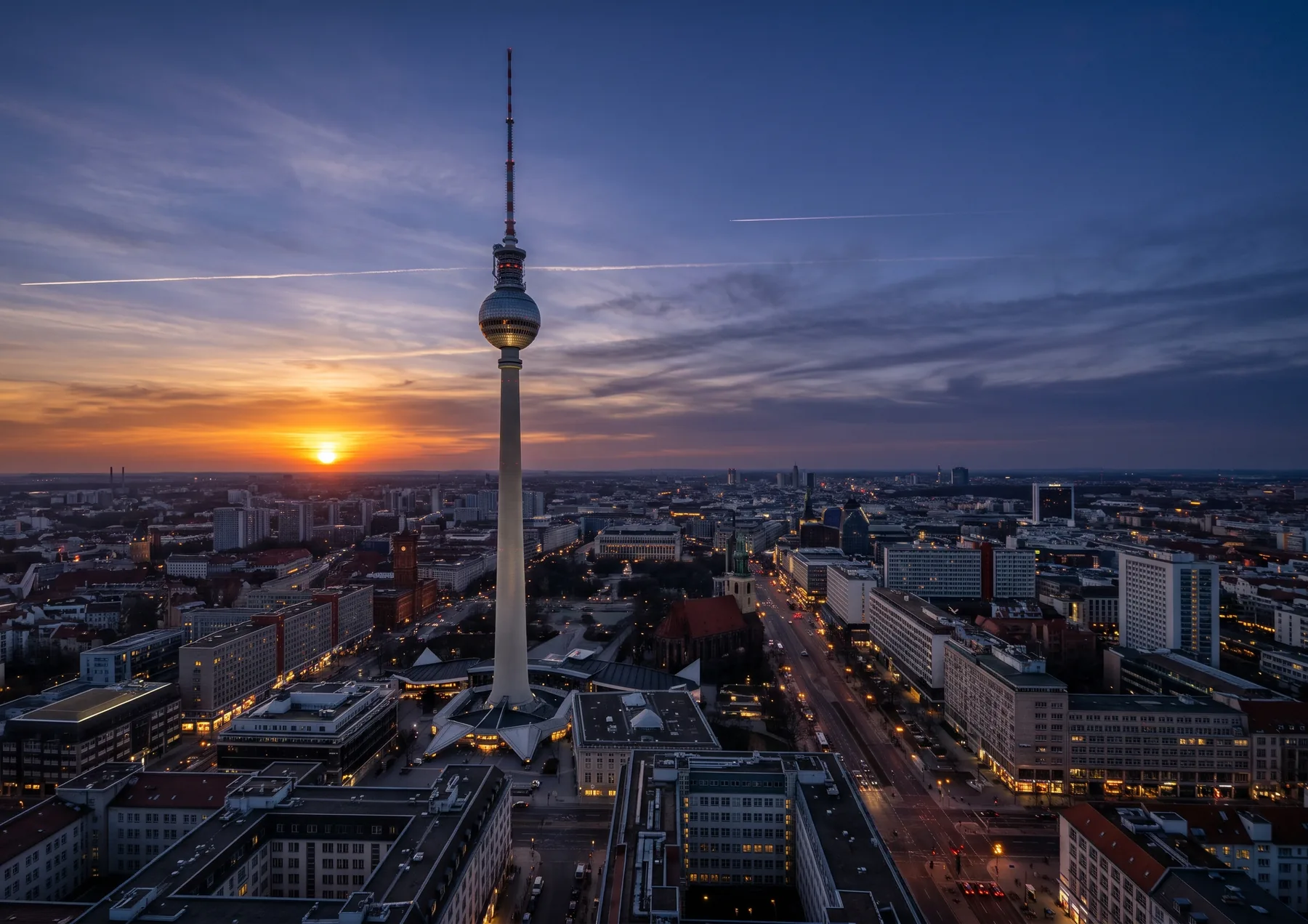 The Berlin television tower over the city at sunset, the sun low on the horizon and the streets lit below.