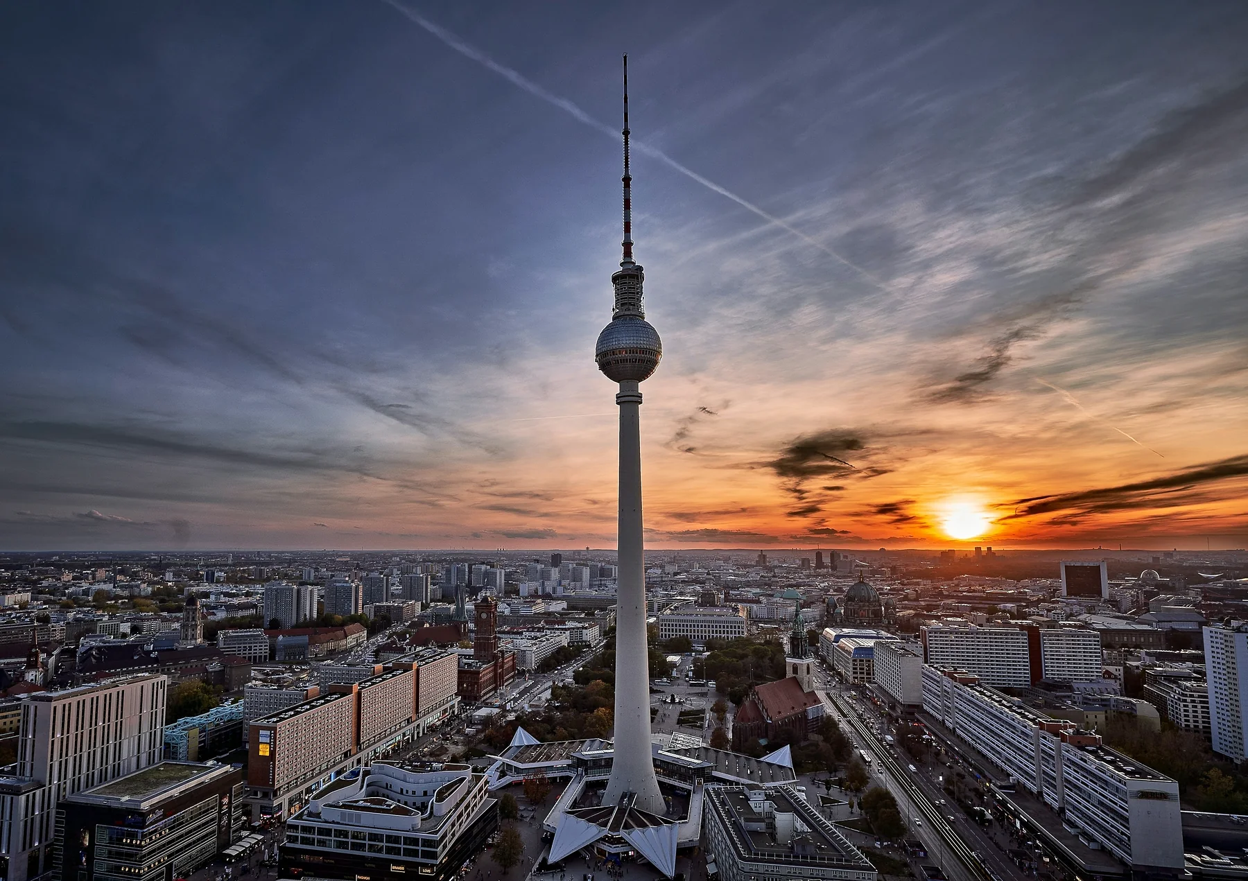 The Berlin television tower standing over the city at sunset, the sky burning orange behind it and the streets already lit below.
